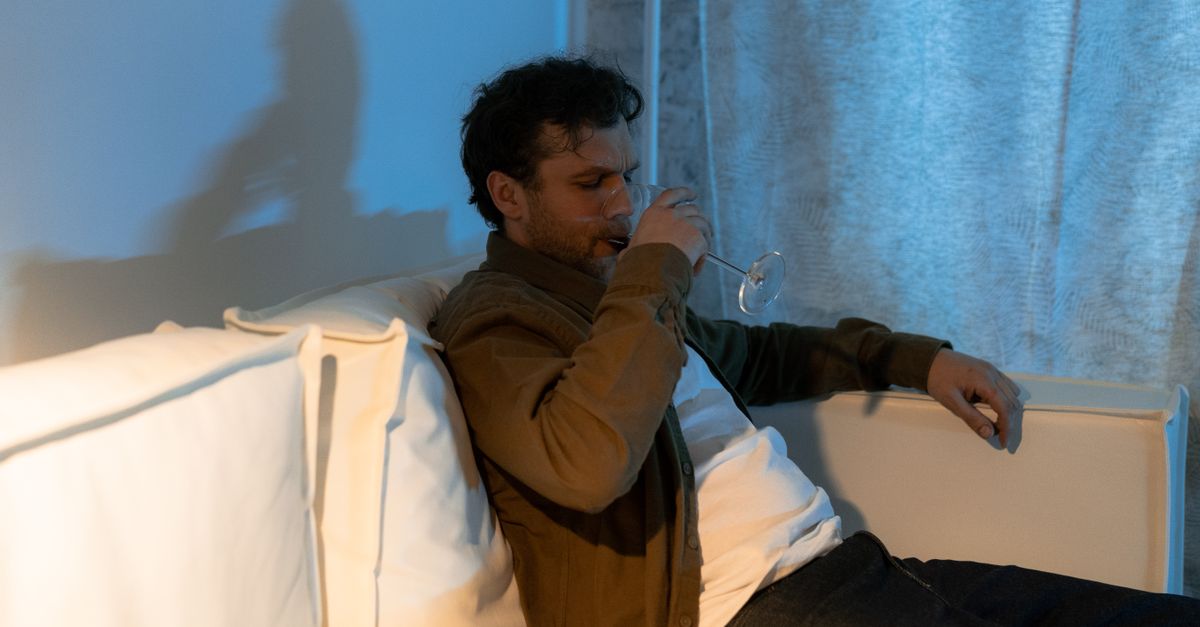 A man sits on a couch, drinking wine in a dimly lit room, conveying a contemplative mood.