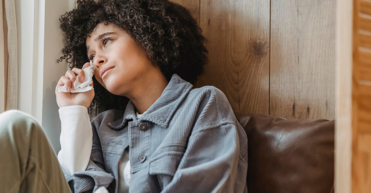Desperate African American female with curly hair wiping face with tissue while crying from sadness on windowsill and looking away