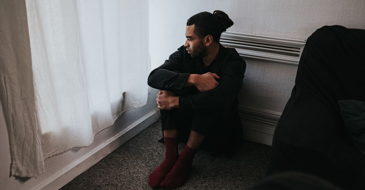 A contemplative man sits in a corner of a dimly lit bedroom, reflecting deep emotions.
