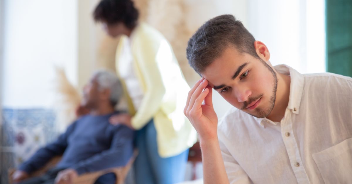 Young man in thoughtful pose indoors with family members in the background, expressing contemplation and care.