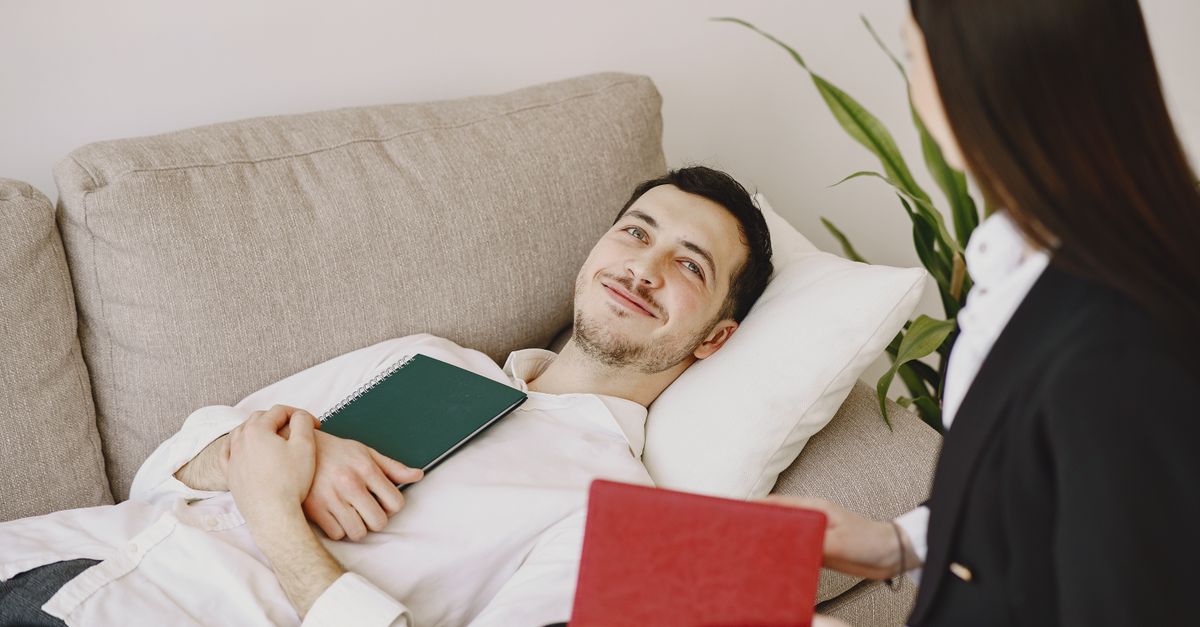 Positive man in formal wear lying on sofa with notebook in hands while smiling and talking with therapist in black suit