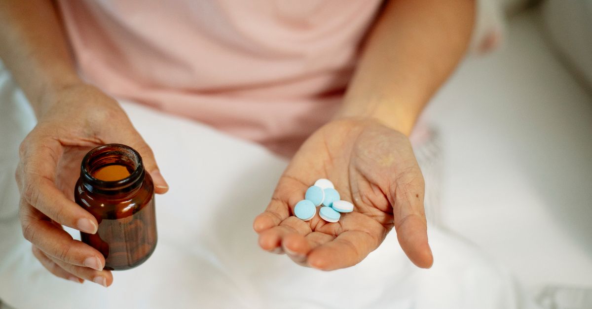 From above of crop unrecognizable person sitting on bed and holding bunch of painkillers in hand at home