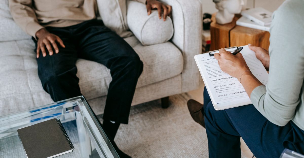 From above crop African American male in casual clothes sitting on comfy couch and answering professional female consultant questions in modern psychotherapy of