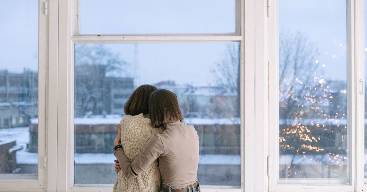 Two people hugging indoors near a window with a snowy, cityscape background.