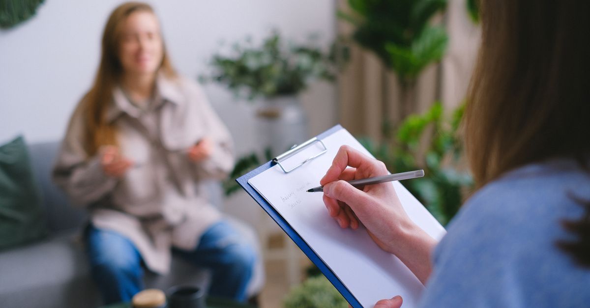 Unrecognizable professional female psychologist writing on clipboard while sitting against client on blurred background during psychotherapy session in light of